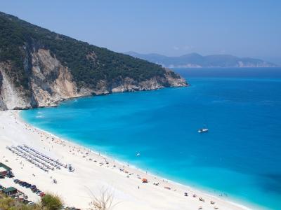 A high-angle view of a wide, white-sand beach with turquoise water, backed by steep, forested cliffs and featuring rows of beach umbrellas and a small parking area.