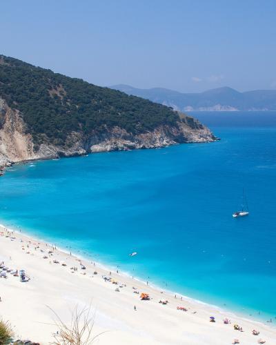 A high-angle view of a wide, white-sand beach with turquoise water, backed by steep, forested cliffs and featuring rows of beach umbrellas and a small parking area.