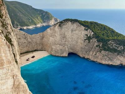 A high-angle view of a secluded cove with steep limestone cliffs, turquoise water, and a rusted shipwreck on the sandy beach.