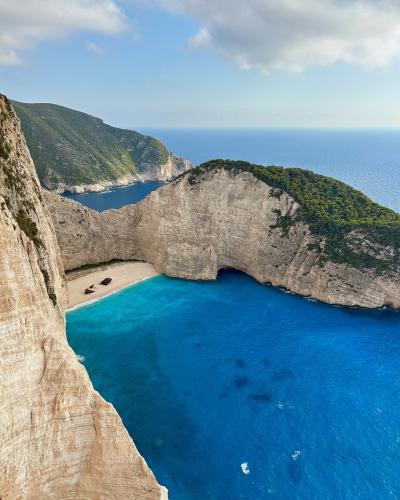 A high-angle view of a secluded cove with steep limestone cliffs, turquoise water, and a rusted shipwreck on the sandy beach.