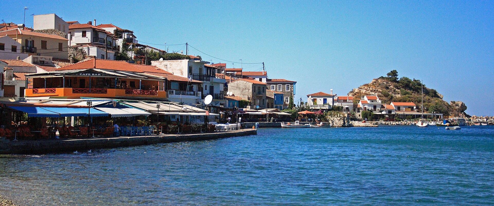 A coastal village scene featuring a waterfront promenade lined with restaurants and cafes, terracotta-roofed buildings climbing a hillside, and a small rocky islet in the bay.