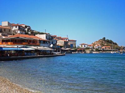A coastal village scene featuring a waterfront promenade lined with restaurants and cafes, terracotta-roofed buildings climbing a hillside, and a small rocky islet in the bay.