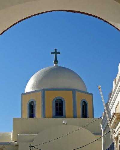A view through a white archway of a yellow and blue Orthodox church dome topped with a cross against a clear blue sky.