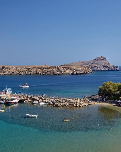 A high-angle view of a sandy beach with rows of sun loungers and umbrellas, a small harbor with several tour boats docked at a stone pier, and a distinctive rocky headland with a small tower in the background.