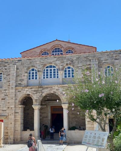 A large, historic stone church with a distinctive facade featuring three arched windows on the upper level and a central arched entrance supported by columns, set against a clear blue sky with tourists present.