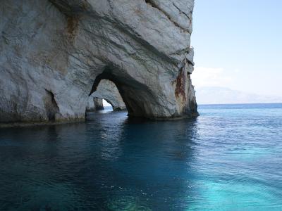 A view from the water looking into a sea cave with a natural archway in a white limestone cliff, with distant mountains visible across the blue sea.