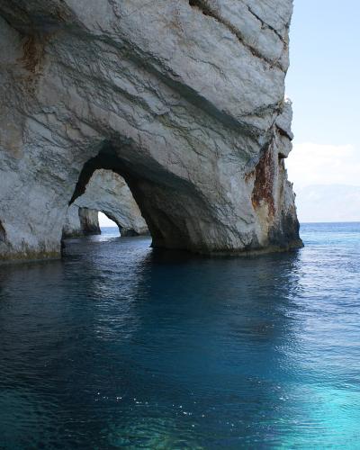 A view from the water looking into a sea cave with a natural archway in a white limestone cliff, with distant mountains visible across the blue sea.