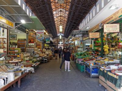 An interior view of a covered market hall with a high, arched metal truss roof, featuring stalls selling food products like cheese, wine, and produce, with Greek signage visible on the shop fronts.