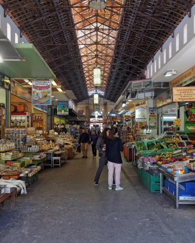 An interior view of a covered market hall with a high, arched metal truss roof, featuring stalls selling food products like cheese, wine, and produce, with Greek signage visible on the shop fronts.