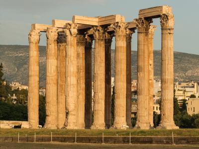 A row of massive, fluted Corinthian columns with ornate capitals, part of a ruined ancient temple, set against a backdrop of a modern city and hills.