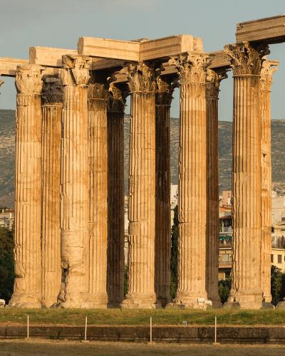 A row of massive, fluted Corinthian columns with ornate capitals, part of a ruined ancient temple, set against a backdrop of a modern city and hills.