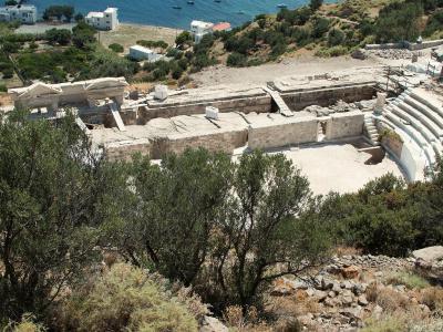 An ancient stone theater with tiered seating and a stage structure, situated on a hillside overlooking the sea with white buildings in the background.