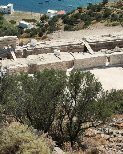 An ancient stone theater with tiered seating and a stage structure, situated on a hillside overlooking the sea with white buildings in the background.