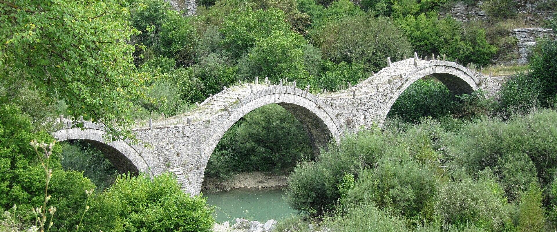 The triple-arched Kalogeriko (Plakidas) stone bridge near Kipi in Zagori, Epirus, spanning a green river among wooded limestone slopes