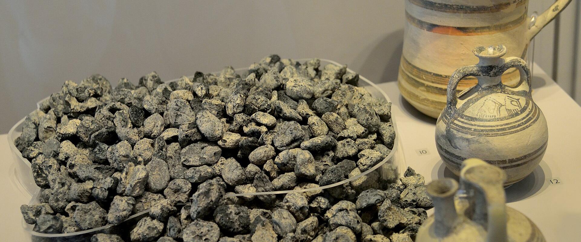 A display case containing a large pile of carbonized organic remains (identified as figs) alongside two small ceramic vessels and one larger ceramic pitcher on a white shelf.