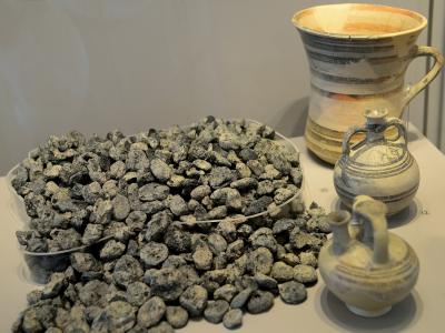 A display case containing a large pile of carbonized organic remains (identified as figs) alongside two small ceramic vessels and one larger ceramic pitcher on a white shelf.