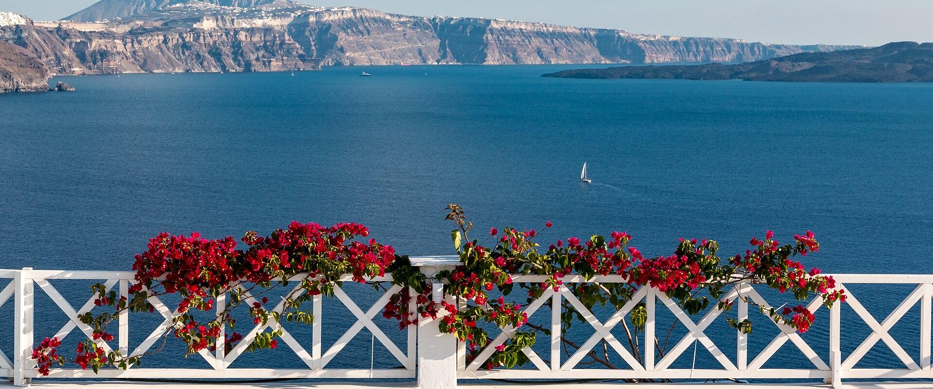 A view from a white terrace with a white lattice railing and red bougainvillea flowers, looking out over the deep blue caldera waters towards the volcanic island cliffs of Santorini.