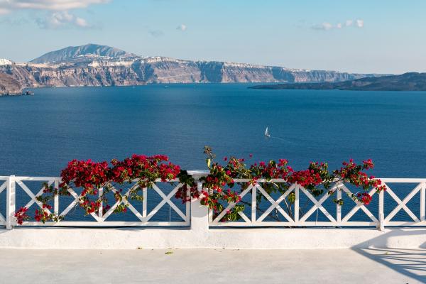 A view from a white terrace with a white lattice railing and red bougainvillea flowers, looking out over the deep blue caldera waters towards the volcanic island cliffs of Santorini.