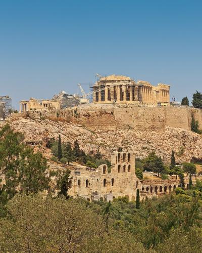 A view of the Acropolis of Athens, featuring the Parthenon and other ancient ruins on a rocky hill, with the Odeon of Herodes Atticus visible below amidst trees.