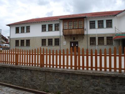 A white, two-story building with a traditional architectural style, featuring a stone base, a central wooden balcony, and a red-tiled roof, situated behind a wooden fence.