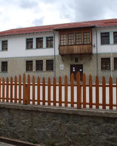 A white, two-story building with a traditional architectural style, featuring a stone base, a central wooden balcony, and a red-tiled roof, situated behind a wooden fence.