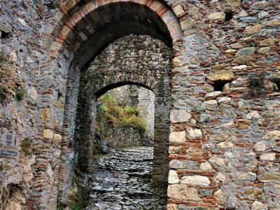 A stone archway with brick voussoirs leading through a cobblestone path, featuring a small white sign on the left wall with Greek text and English translation "PALACE ENTRANCE" and "MONEMVASIA GATE".