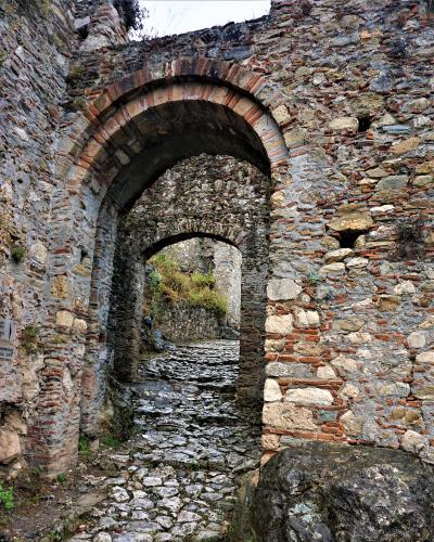 A stone archway with brick voussoirs leading through a cobblestone path, featuring a small white sign on the left wall with Greek text and English translation "PALACE ENTRANCE" and "MONEMVASIA GATE".
