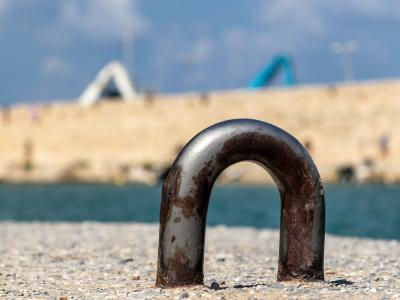 A close-up of a rusty metal mooring bollard on a pebbled quay, with a blurred stone breakwater and blue sky in the background.