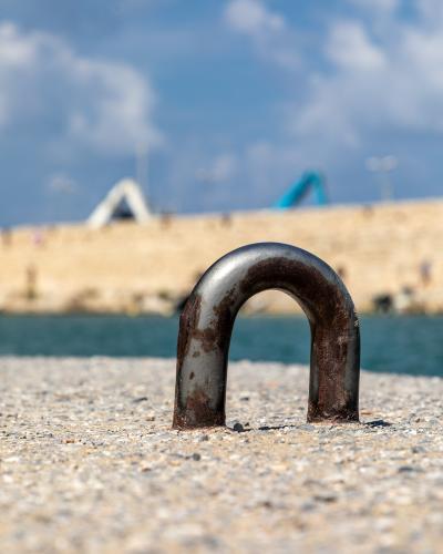 A close-up of a rusty metal mooring bollard on a pebbled quay, with a blurred stone breakwater and blue sky in the background.