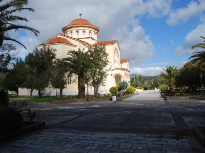 A cream-colored Orthodox church with a red-tiled dome and cross, surrounded by palm trees and a paved courtyard under a cloudy sky.
