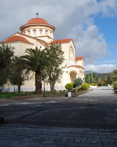 A cream-colored Orthodox church with a red-tiled dome and cross, surrounded by palm trees and a paved courtyard under a cloudy sky.