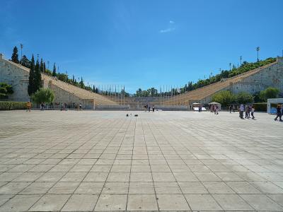 A wide-angle view of a large, U-shaped marble stadium with tiered seating rising on both sides, a paved plaza in the foreground, and a small kiosk labeled "THE OFFICIAL MUSEUM SHOP" on the right.