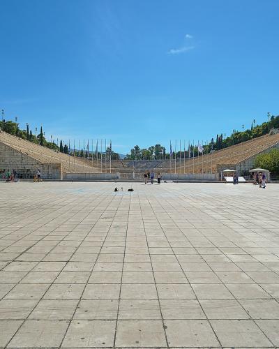 A wide-angle view of a large, U-shaped marble stadium with tiered seating rising on both sides, a paved plaza in the foreground, and a small kiosk labeled "THE OFFICIAL MUSEUM SHOP" on the right.