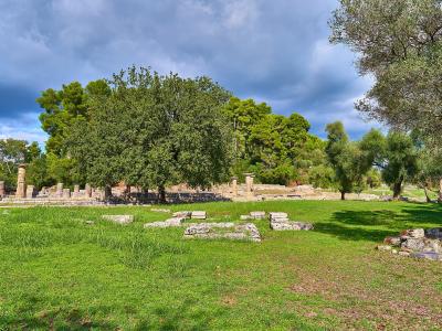 Ancient stone ruins consisting of a low foundation wall and several standing columns set within a grassy field bordered by trees under a cloudy sky.