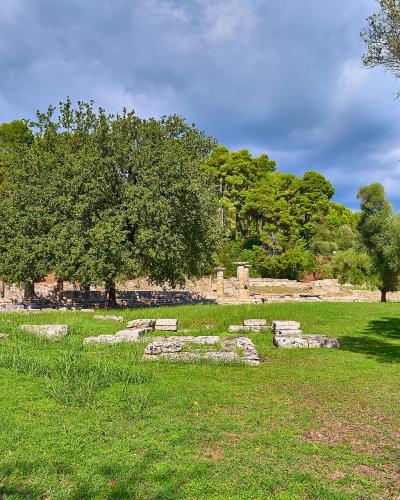 Ancient stone ruins consisting of a low foundation wall and several standing columns set within a grassy field bordered by trees under a cloudy sky.