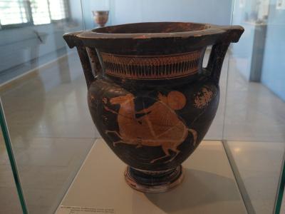 A black-figure Greek krater depicting a mounted warrior, displayed inside a glass museum case with a descriptive label at the base.