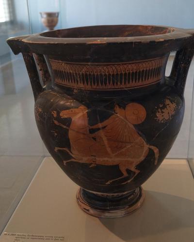 A black-figure Greek krater depicting a mounted warrior, displayed inside a glass museum case with a descriptive label at the base.