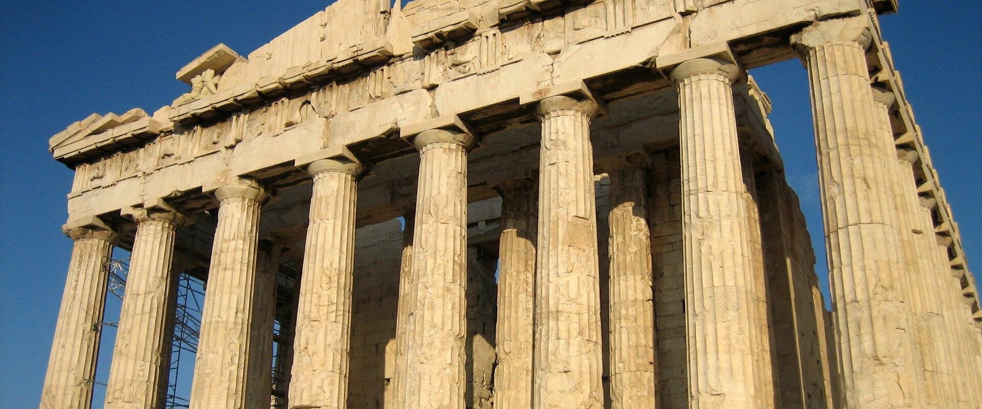 The image shows a low-angle, close-up view of the upper section of a large, ancient marble temple, specifically focusing on the corner where the architrave meets the columns, with a pile of rubble in the foreground.