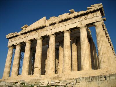 The image shows a low-angle, close-up view of the upper section of a large, ancient marble temple, specifically focusing on the corner where the architrave meets the columns, with a pile of rubble in the foreground.