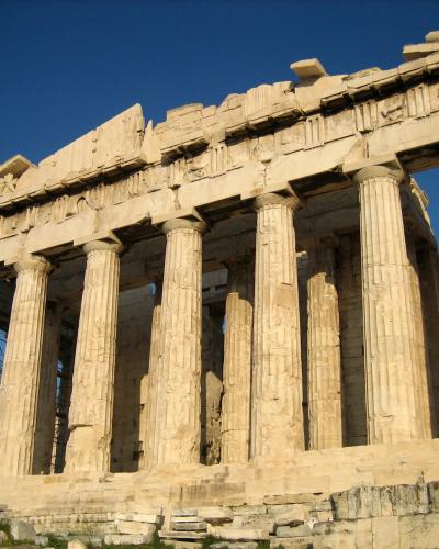 The image shows a low-angle, close-up view of the upper section of a large, ancient marble temple, specifically focusing on the corner where the architrave meets the columns, with a pile of rubble in the foreground.
