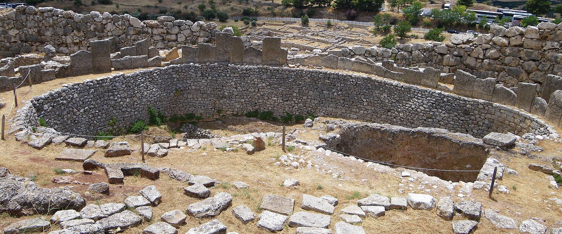 The image shows the circular stone foundations and ruins of the Mycenaean megaron at the Palace of Nestor in Pylos, Greece, with a view of the surrounding landscape and mountains in the background.