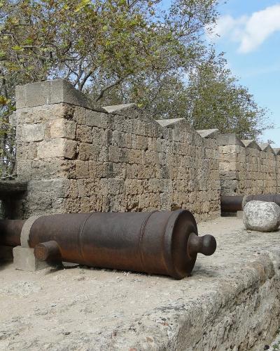A row of old, rusted cannons lined up on a stone parapet next to a crenellated stone wall and a tall stone tower.