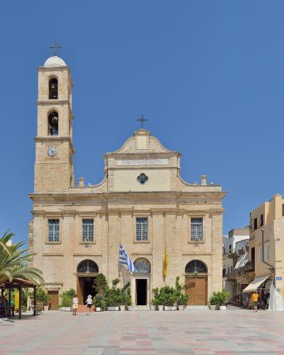 A large, light-stone Orthodox church with a tall bell tower and a Greek flag flying in front, situated in a paved public square with palm trees and surrounding buildings.