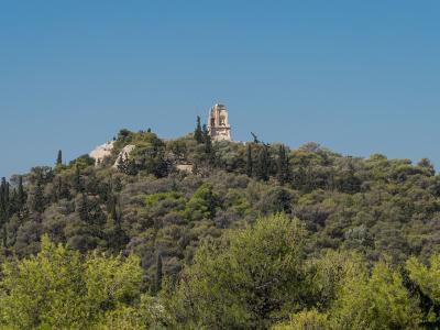 A forested hilltop featuring a prominent ancient stone monument (the Monument of Philopappos) and a bronze statue against a clear blue sky.