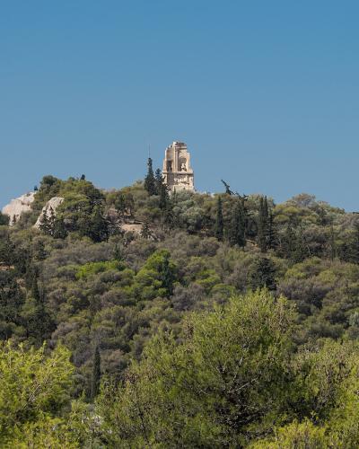 A forested hilltop featuring a prominent ancient stone monument (the Monument of Philopappos) and a bronze statue against a clear blue sky.