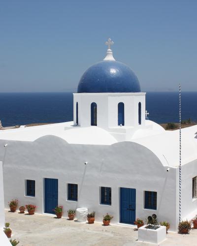 A white-washed church with a prominent blue dome and a separate bell tower, situated on a cliff overlooking the deep blue sea under a clear sky.