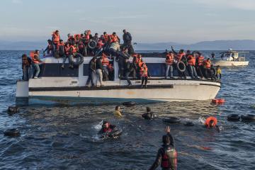 A heavily overcrowded white motorboat filled with refugees wearing orange life vests is shown on the water, with several people in the sea nearby and a smaller boat in the background.