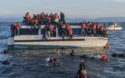 A heavily overcrowded white motorboat filled with refugees wearing orange life vests is shown on the water, with several people in the sea nearby and a smaller boat in the background.