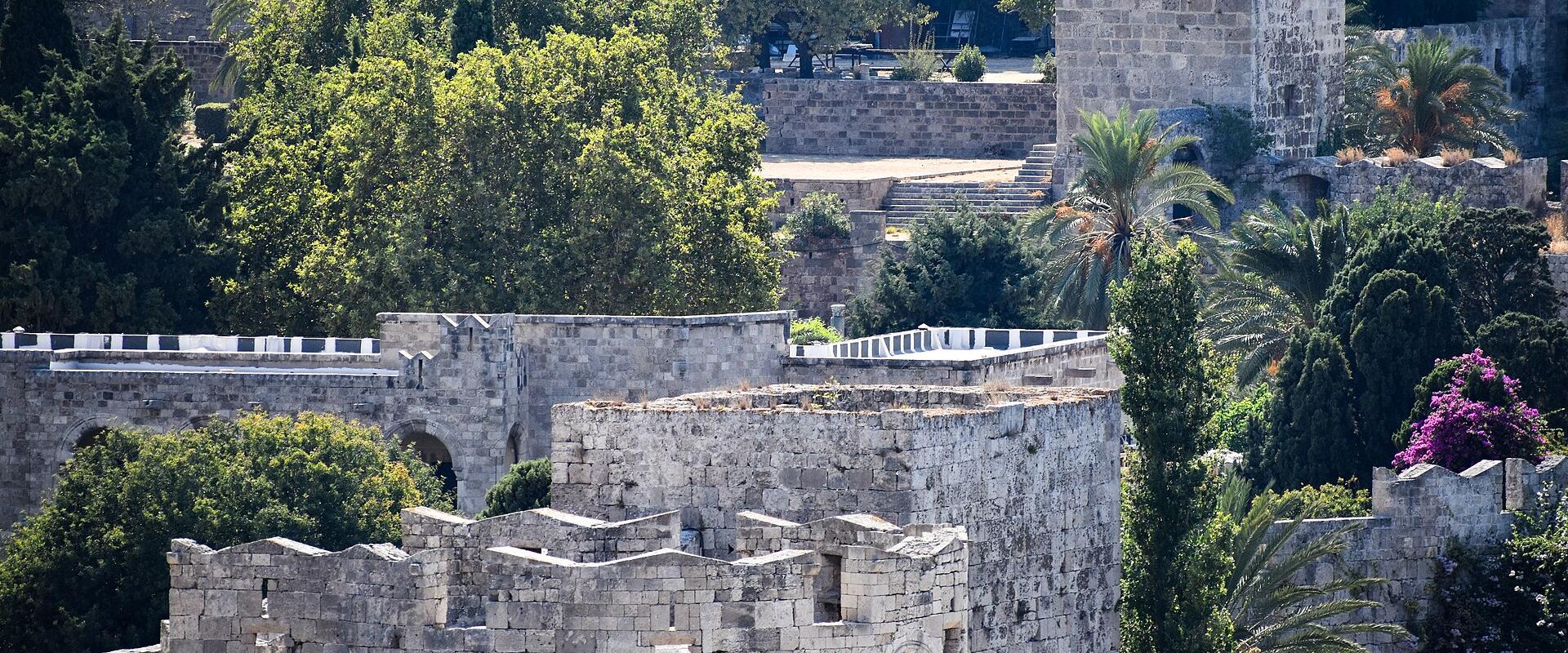 The image shows stone fortifications with crenellated walls and towers, interspersed with lush green trees and vegetation, including palm trees and flowering bushes.