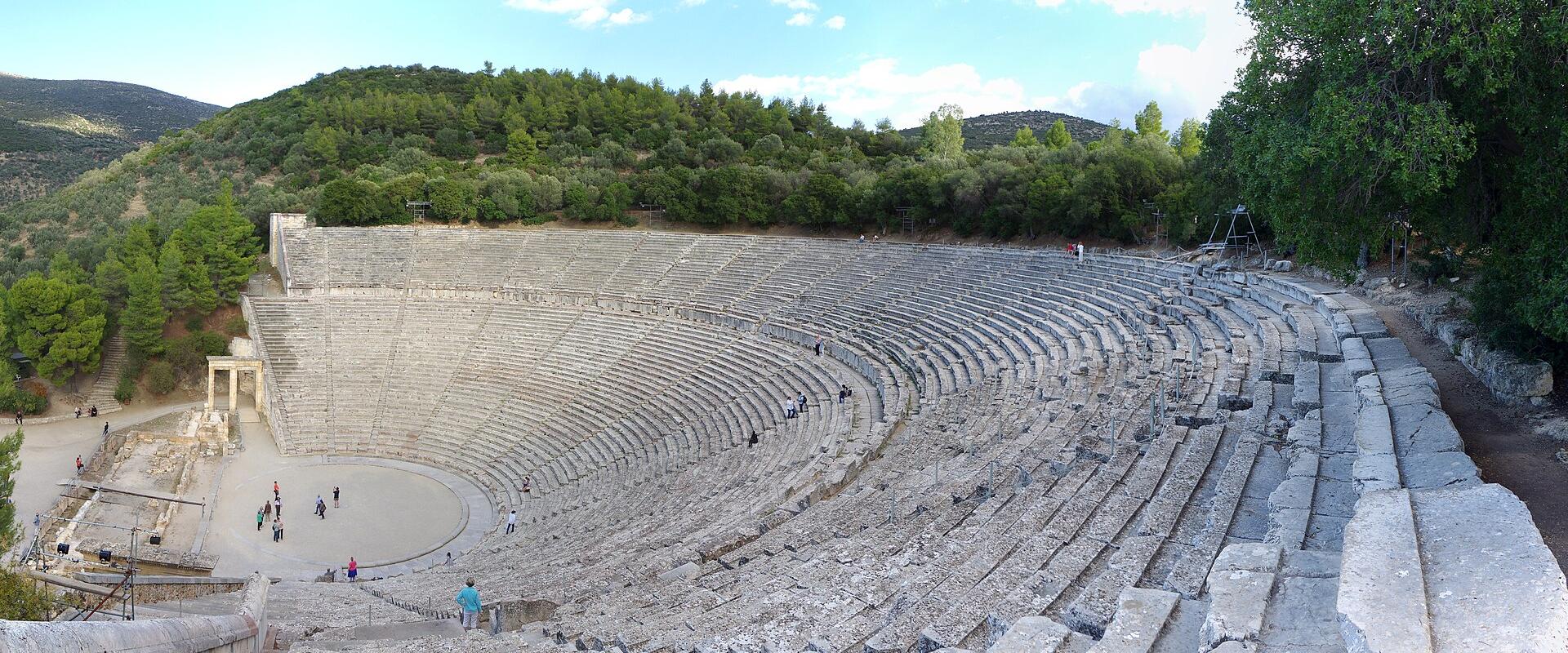 A wide-angle view of the ancient stone seating of a large Greek theater with a circular orchestra and a small temple-like structure (the Thymele) on the left, set against a backdrop of green hills.